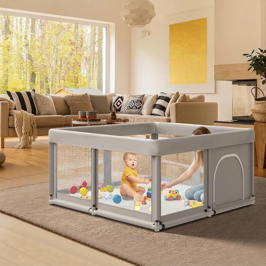 Back-yard scene mid-assembly: parent snapping the last connector on the grey 50×50 Fshibila playpen, picnic blanket beside a cooler, baby crawling toward the open frame